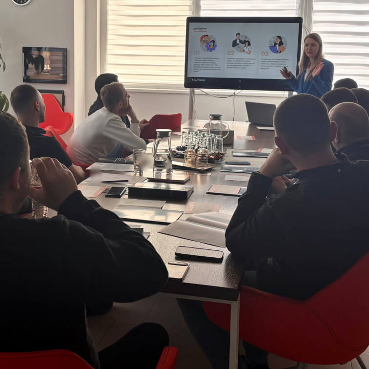 a woman standing in front of a large table with people sitting around it
